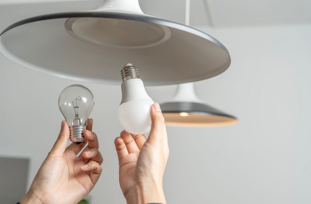 A LED lightbulb and a regular lightbulb being help up to an overhead lamp on the ceiling.