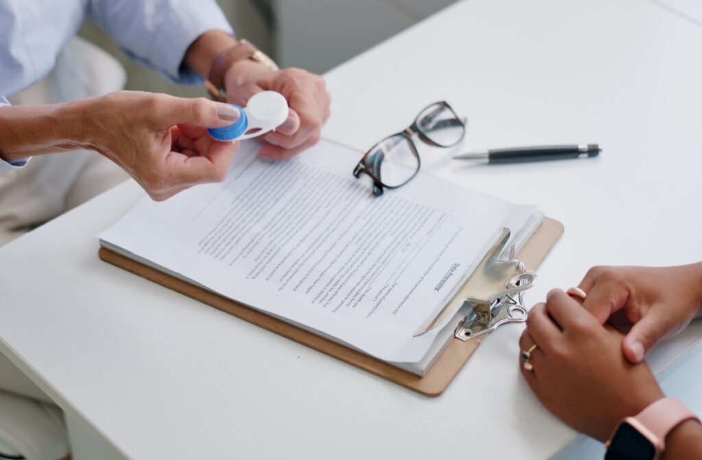 An optometrist holds out a pair of contact lenses to a patient over a clipboard of information about proper contact lens care