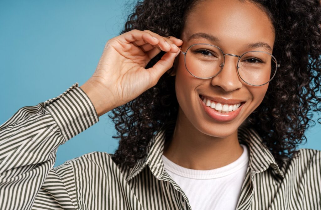 A close up of a person smiling, holding their glasses with one hand.